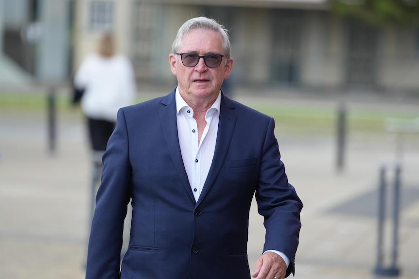 A man in a blue suit, white shirt and sunglasses walking outside.
