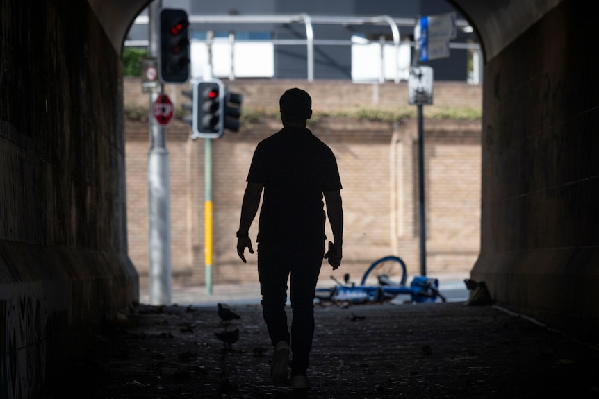 John wearing blue jeans and a black shirt walking through a tunnel towards a street.