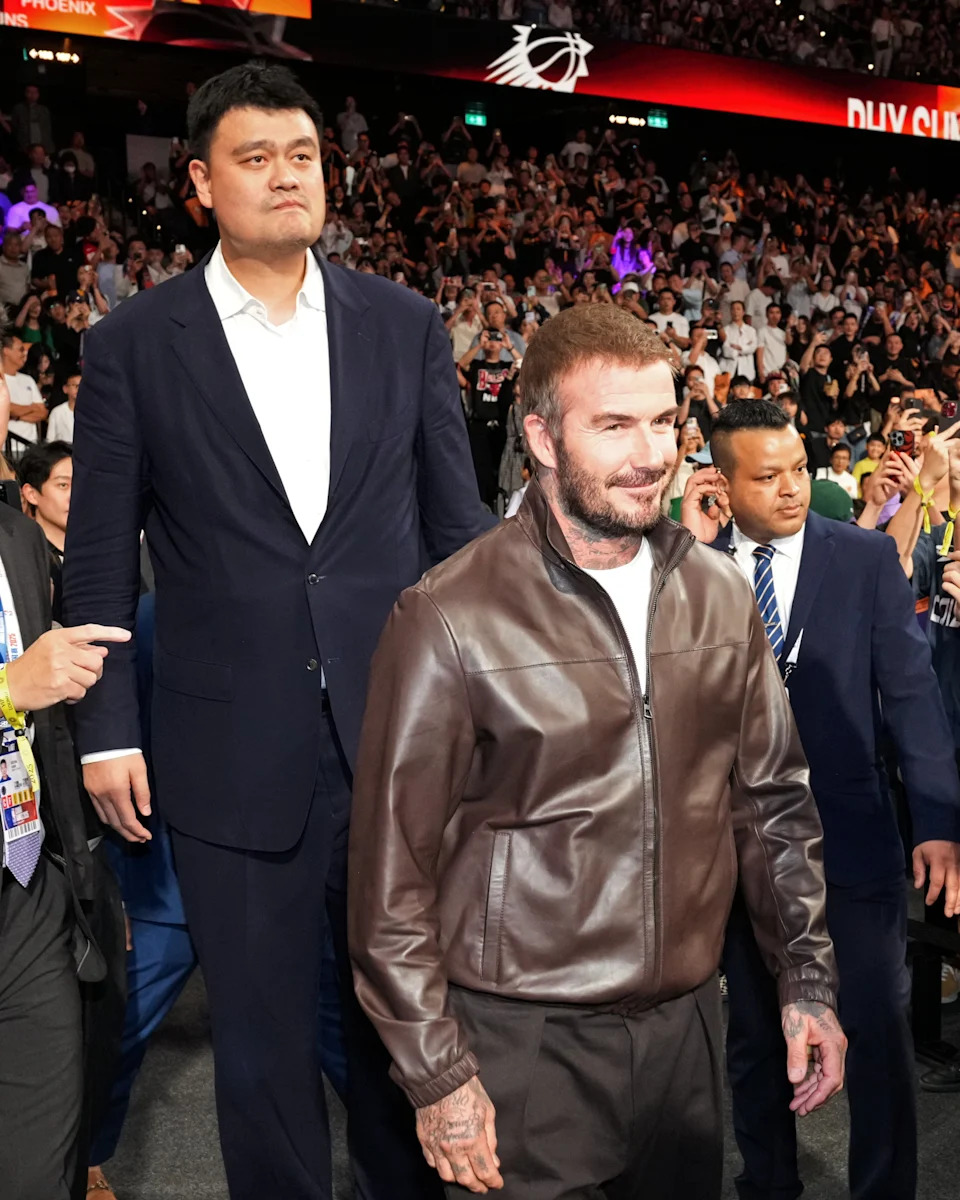 Two well-dressed men at a sports event, one in a suit and the other in a leather jacket, walking through a crowded venue