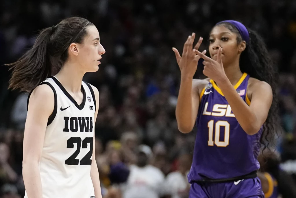 Two female basketball players face each other on the court, one in a white Iowa jersey and the other in a purple LSU jersey, ready for play.