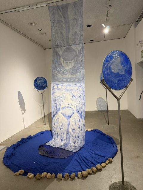Mixed-media installation featuring blue textile elements, circular forms, and natural materials at Heavy Metal.