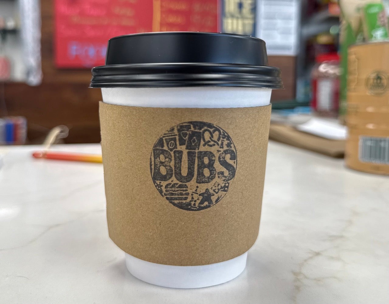 A photo of a small white disposable coffee cup with a black lid with a cardboard sleeve that reads BUBS on it.