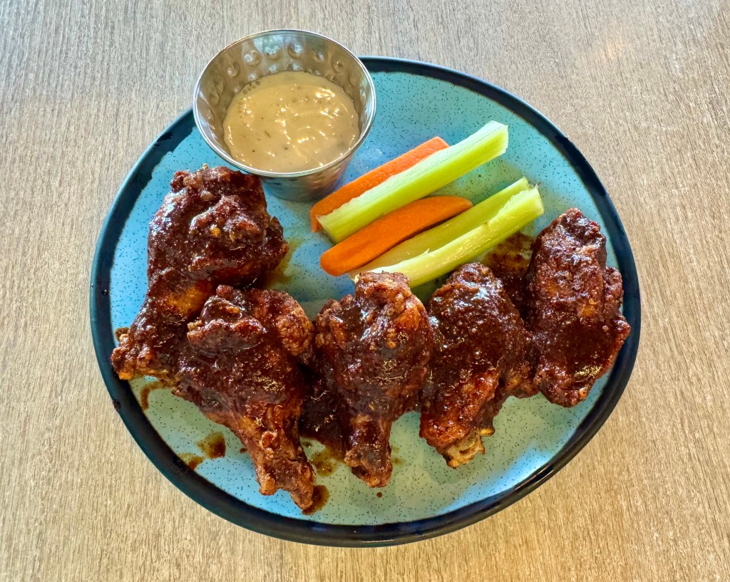 Five wings tossed in aguachile negro on a plate with ranch dressing and crudites.