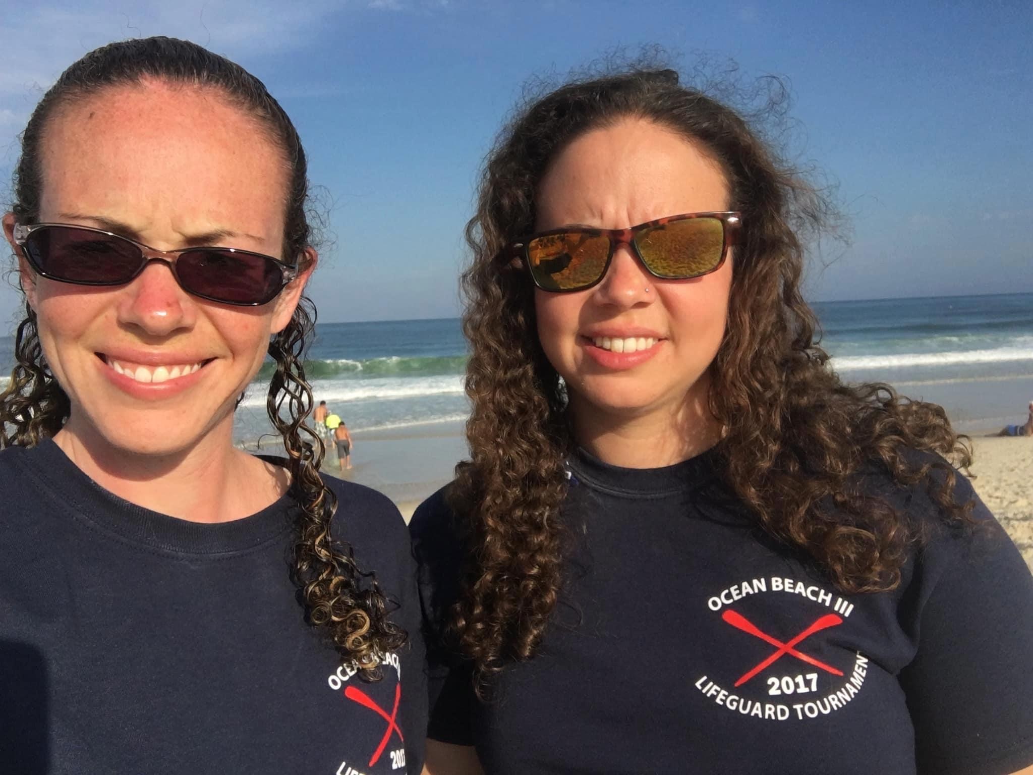 Emily Hoffman and Beth Hoffman stand together on a beach in Ocean Beach, New Jersey, in 2017, wearing sunglasses and matching dark shirts from a lifeguard tournament. Beth is on the left and Emily is on the right.