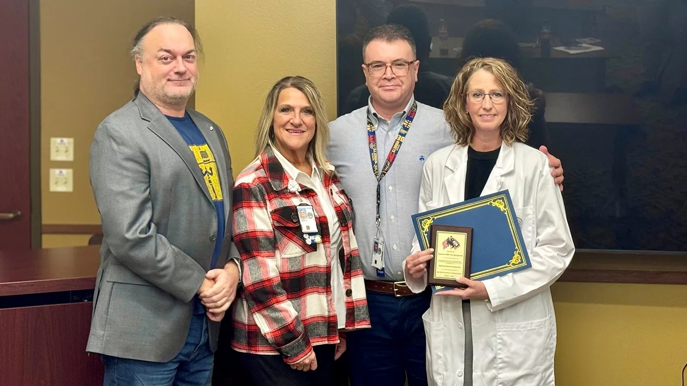 A group of individuals stands together in a meeting room as one person in a white coat holds a framed certificate, recognizing contributions to pain management efforts.