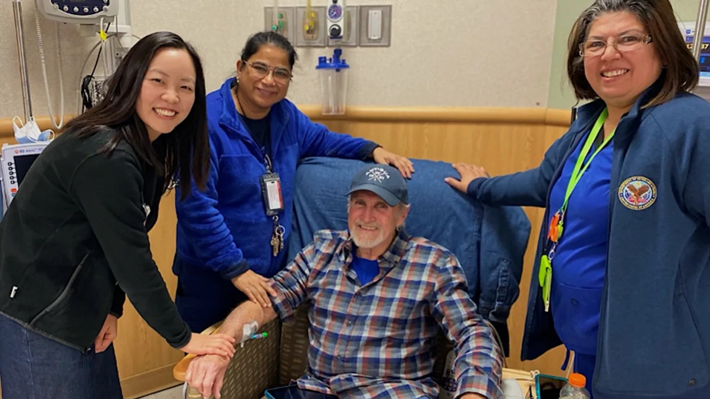 A person sits in a medical chair receiving an IV while three healthcare staff members stand nearby with hands resting on the patient’s arm and chair, representing support during cancer treatment.