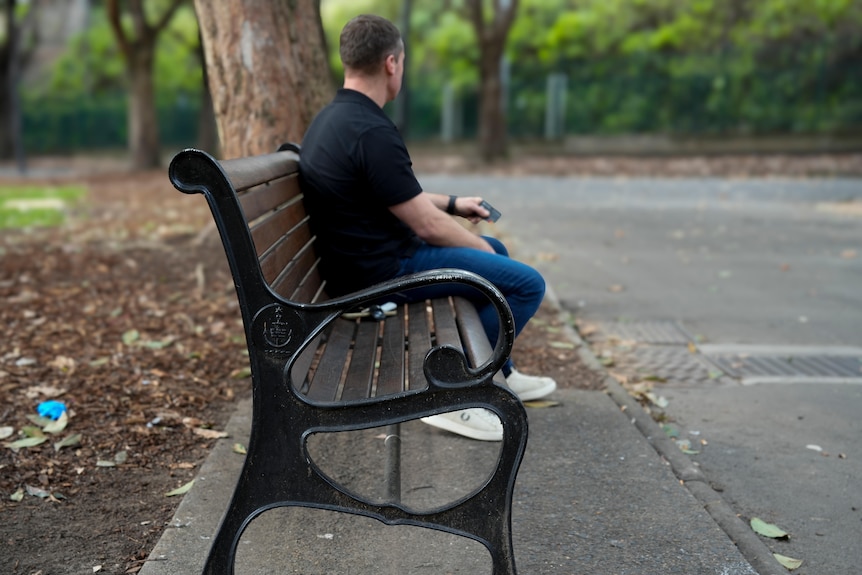 John wearing a black shirt and jeans sits on a wooden bench at a park looking away from the camera.