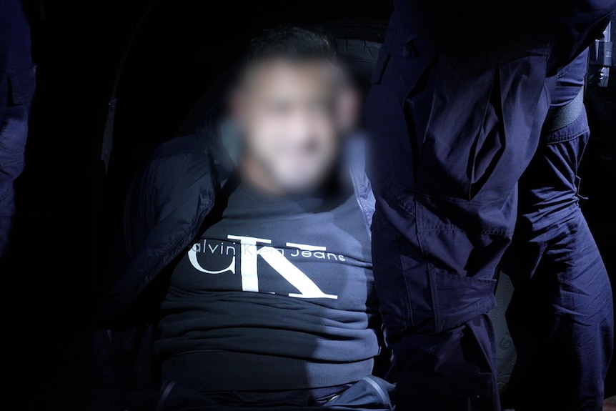 Man in black tracksuit, surrounded by police. Faces blurred