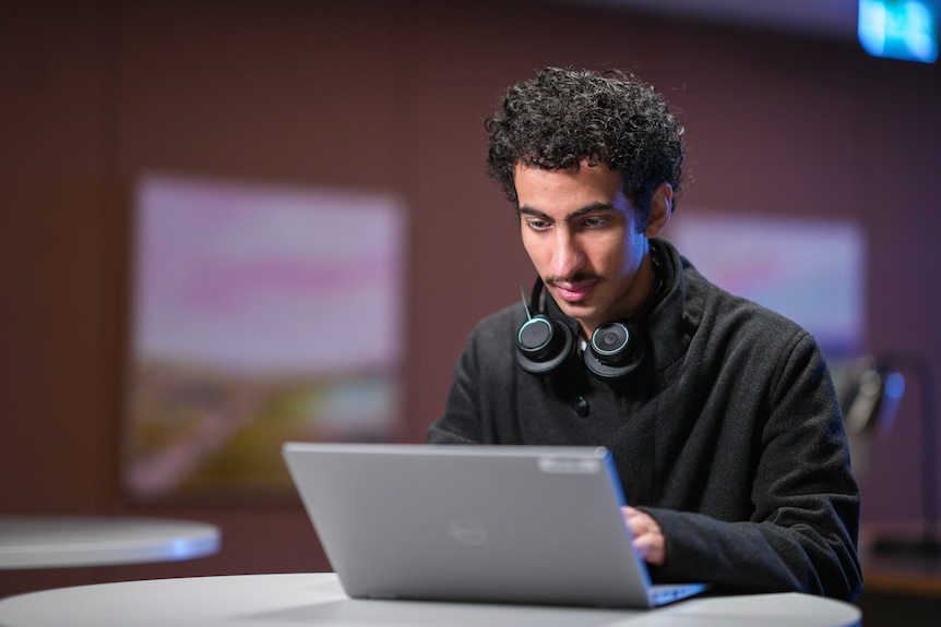 A young man with a moustache and dark, curly hair sits at a table and uses a laptop.