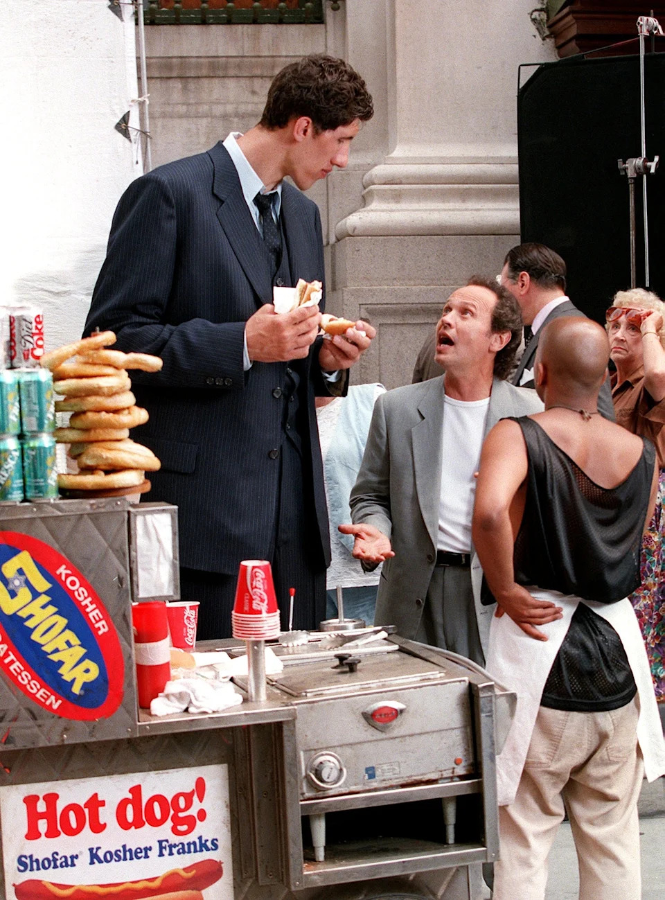 Tall man in a suit eats a hot dog at a food cart while talking to a shorter man gesturing animatedly in a casual gray blazer