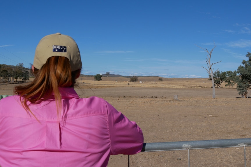 A woman in a pink shirt and cap looking out over a brown paddock.