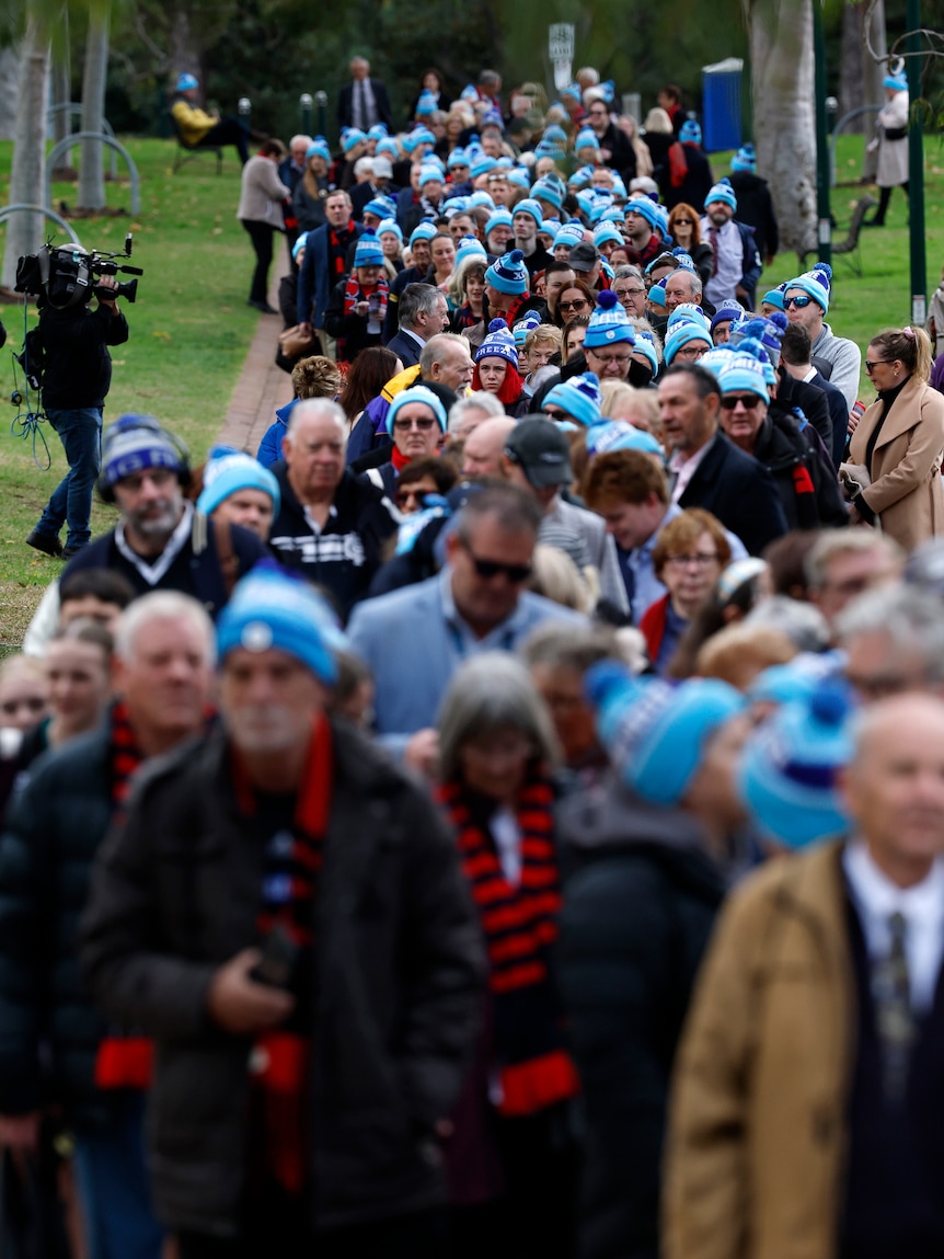 A large line of people stretches back to the rear of the shot, with most people wearing a sky blue beanie.