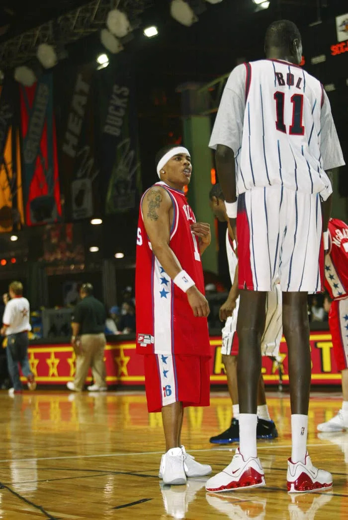 Two basketball players on the court during a casual sports event, wearing loose jerseys and shorts. One player is notably taller