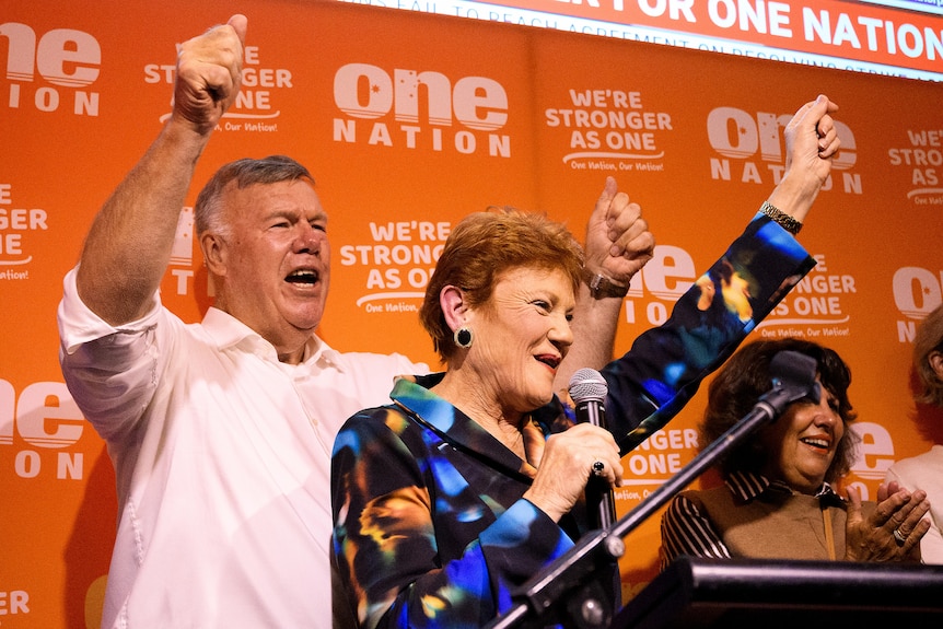 Man and woman pump their fists on stage before a crowd in front of an orange One Nation poster
