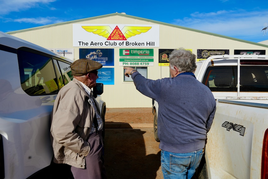 a man with a hat and another man with a blue jumper pointing to a shed with signs on it