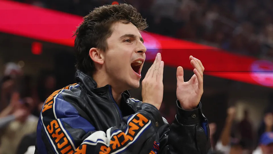 Timothee Chalamet in a Knicks jacket cheering at a sports event, with a red banner in the background.