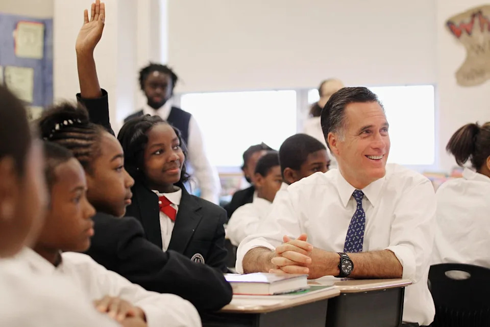 Man in white dress shirt and tie sits at school desk around middle school students in uniforms