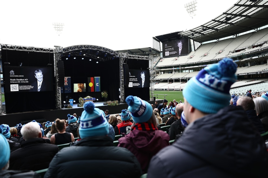 People wearing blue beanies sit in rows of a sports stadium looking towards a stage which has a coffin, flags and flowers.