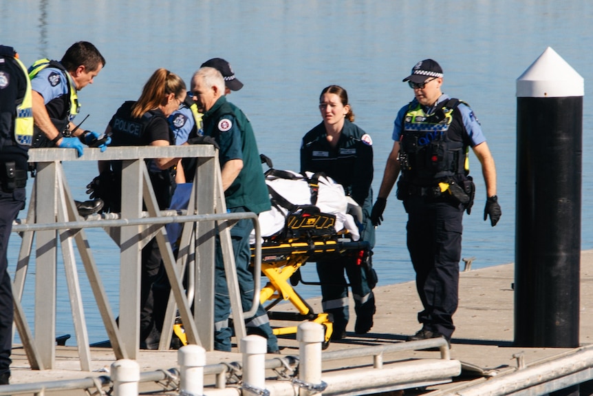 Police and paramedics walk with a person on a stretcher being wheeled along a jetty.