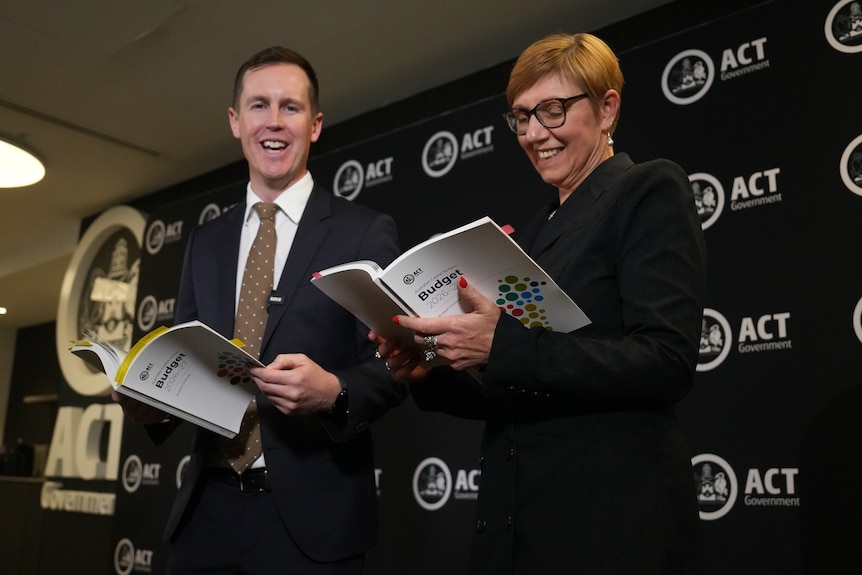A man and woman in suits hold budget papers while smiling.