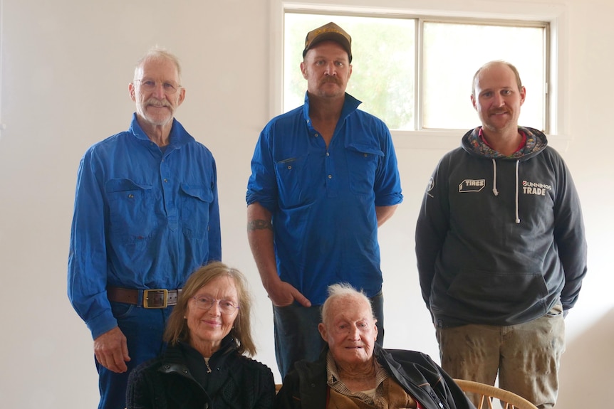 three men standing, two with blue shirts and one with a grey jumper, a lady with glasses sitting next to an old man