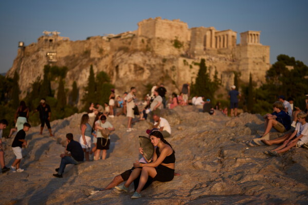 A tourist uses a hand fan to cool off during a heat wave as people gather on Areopagus Hill, with the Acropolis in the background, in Athens, Tuesday, July 21, 2026. (AP Photo/Petros Giannakouris)