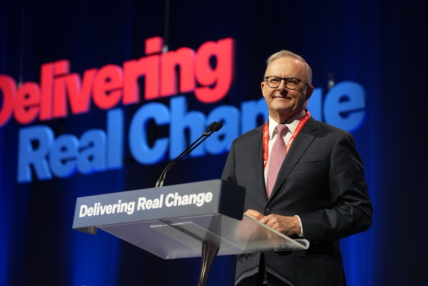 Anthony Albanese smiles while standing at a podium. The words "Delivering Real Change" appear on the podium and on the wall