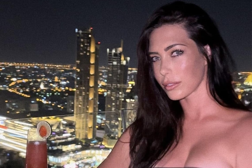 a brunette woman with a city behind her at night. A drink sits on the table next to her. 
