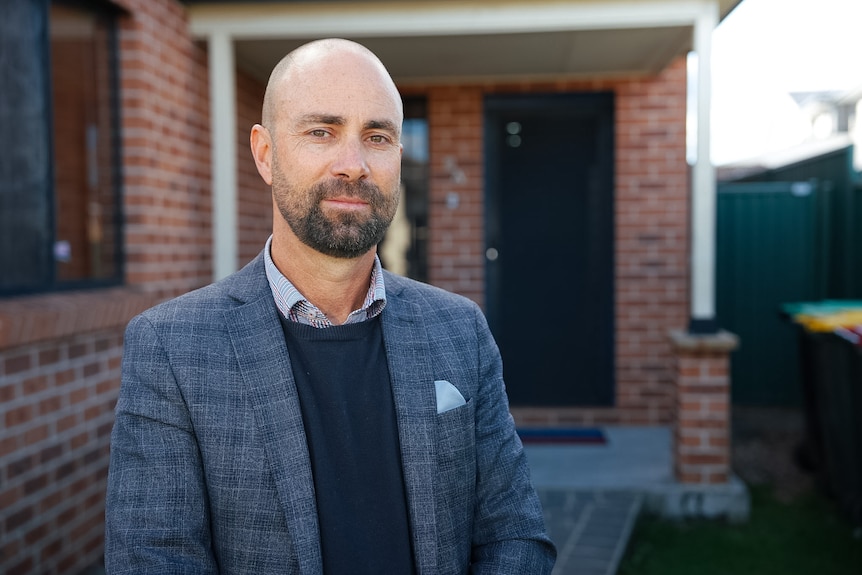 Mr Attard wearing a chequered blazer, blue knit sweater and collared shirt smiles in front of a house.