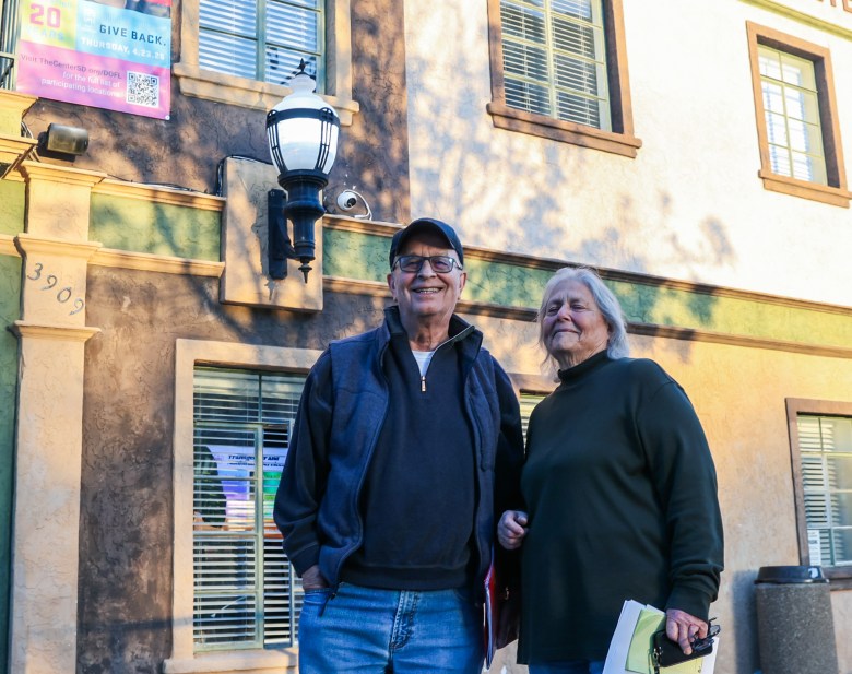 Two seniors in jeans and longsleeves outside a stucco building