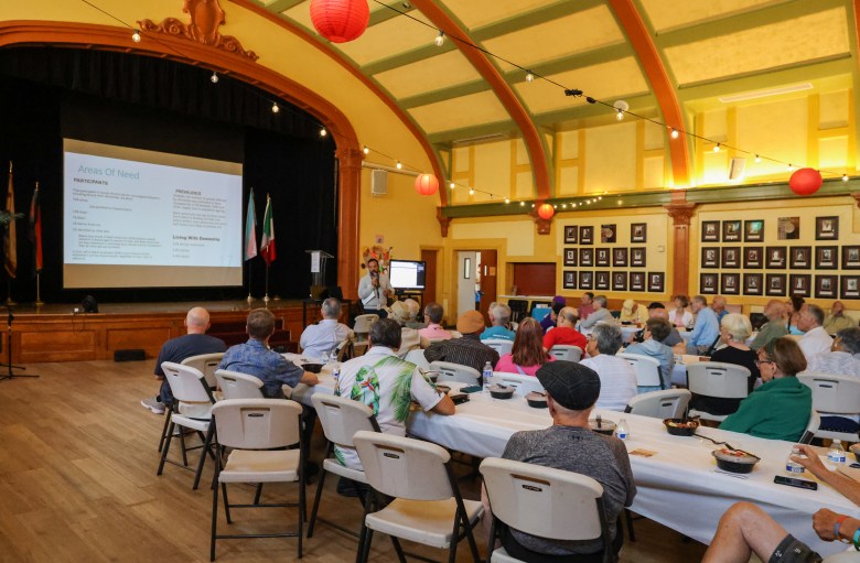 tables of people sitting in large auditorium with hardwood floors, a stage and arched ceiling.