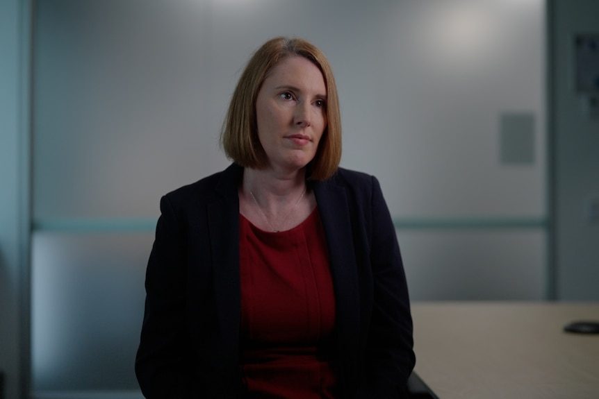 A woman in a dark blazer sits in an office, looking to the side with a serious expression.