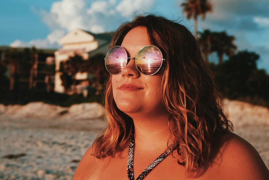 A smiling woman wearing sun glasses at the beach looks off into the distance.