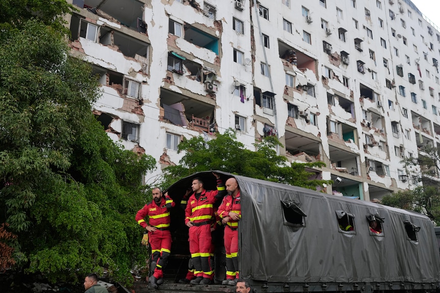 Three men in red and yellow protective jumpsuits standing at a black tent tunnel, in front of a damaged white building.