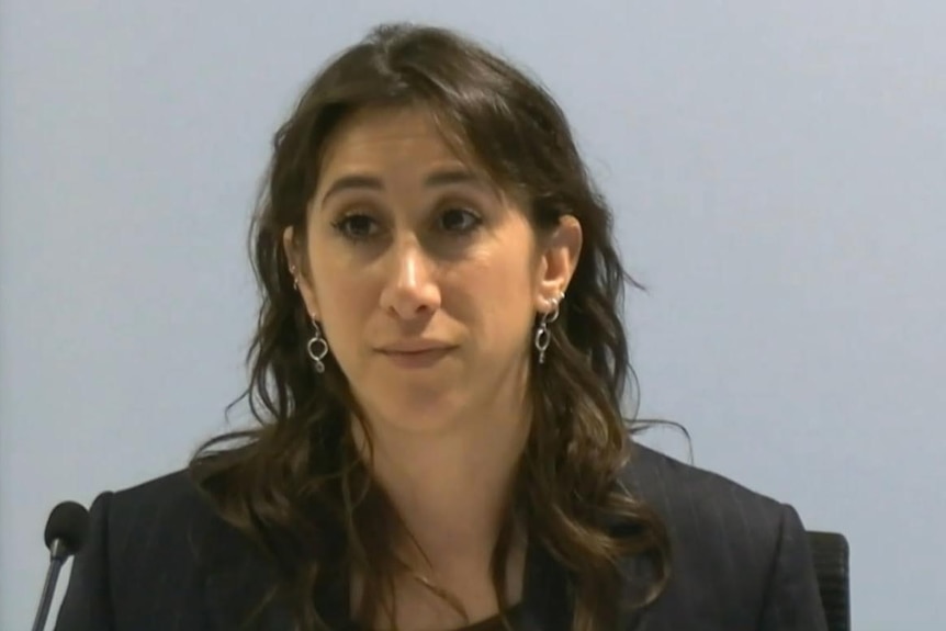 Ms Schwartz sitting at a table behind a microphone while looking serious and wearing a black blazer.