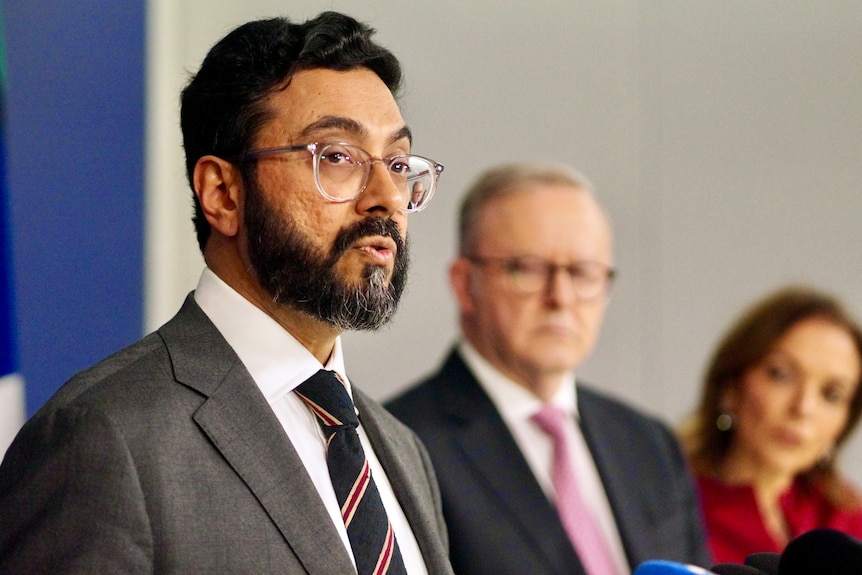 A man wearing a grey suit and spectacles talks, with the prime minsiter in the background