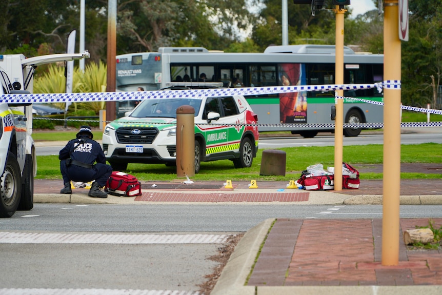 A police officer kneels down next to a police vehicle at a crime scene marked by police tape at an intersection.