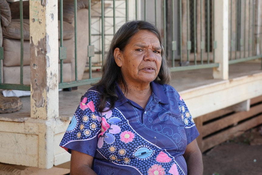 An Aboriginal grandma sits in a colourful shirt in front of a porch