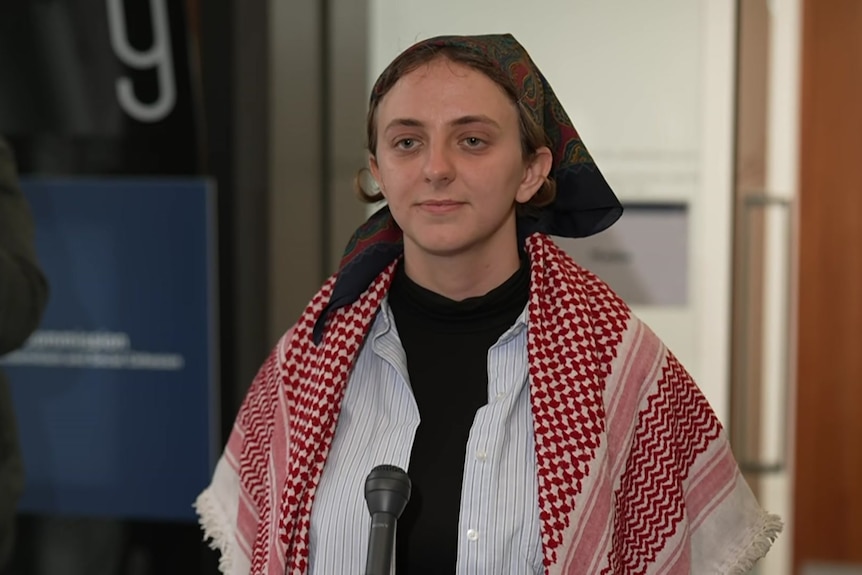 A woman in a brown patterned head scarf, a white shirt over a black top and a red and white checked scarf around her shoulders.