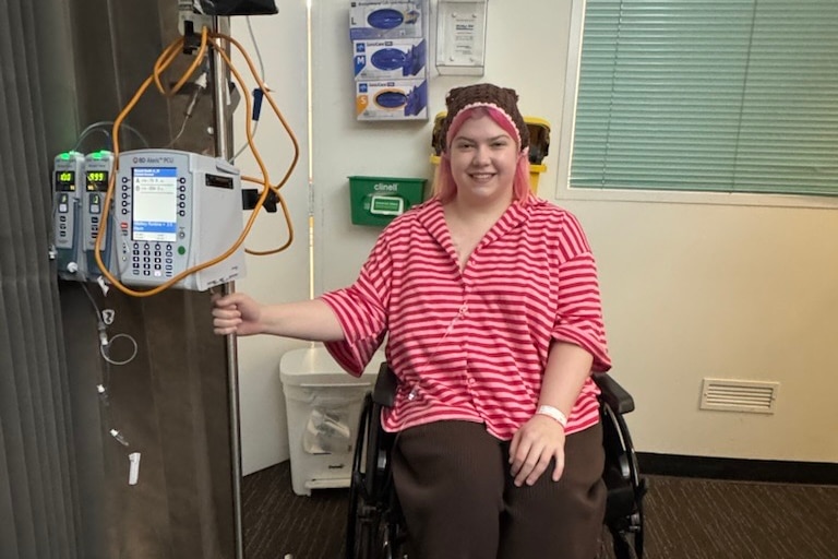 A young woman in pink striped pyjamas, holds onto a machine giving her treatment as she sits smiling in her wheelchair.