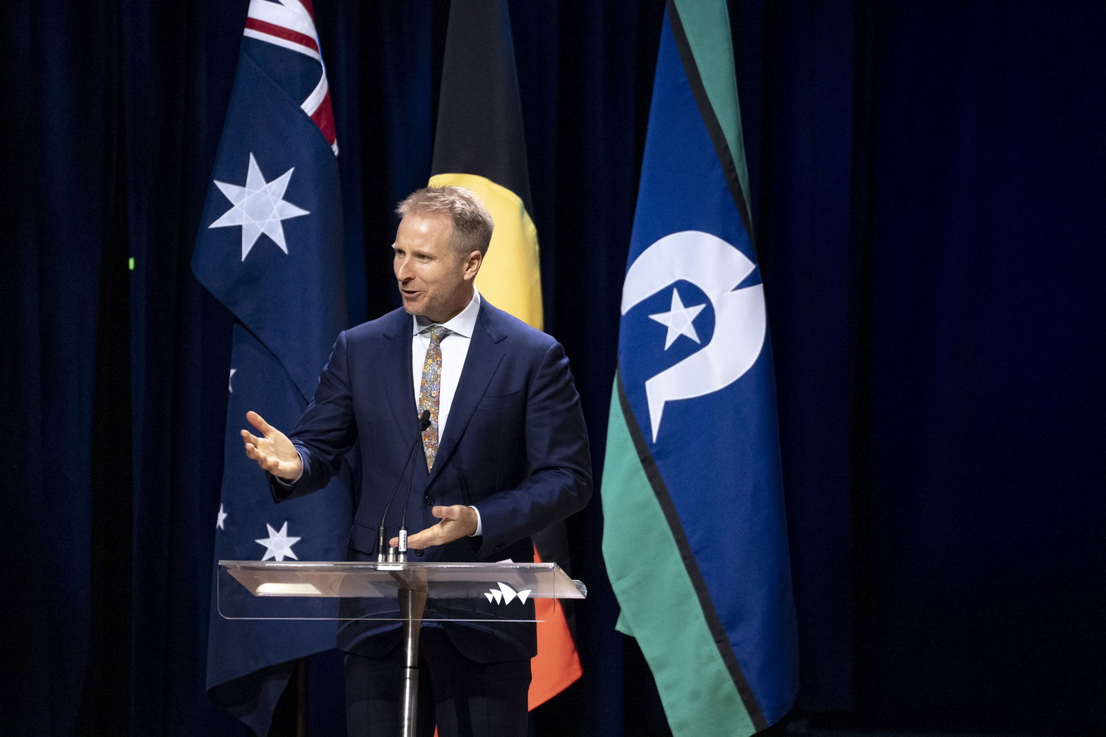 Hamish Macdonald in a navy suit speaking in front of Australian and Indigenous flags.