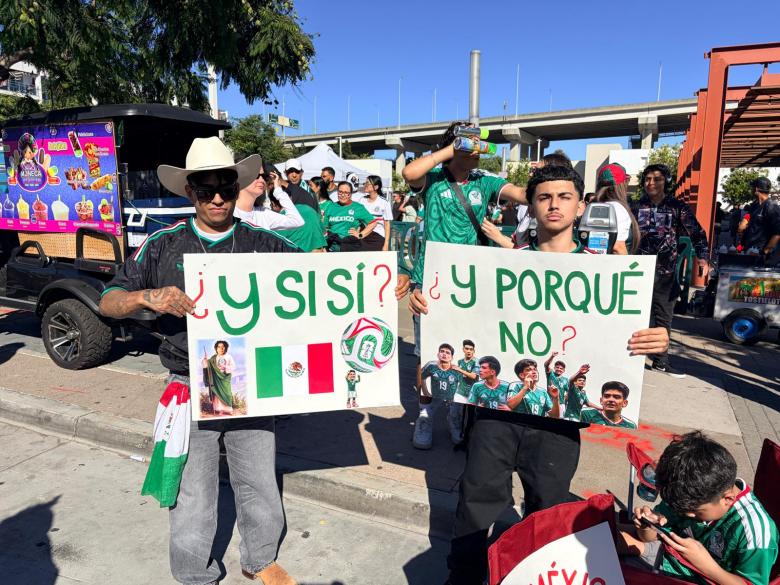 Two men holding signs with Spanish text at a sports event. One sign features a Mexican flag and soccer imagery. Crowd in green jerseys celebrates.