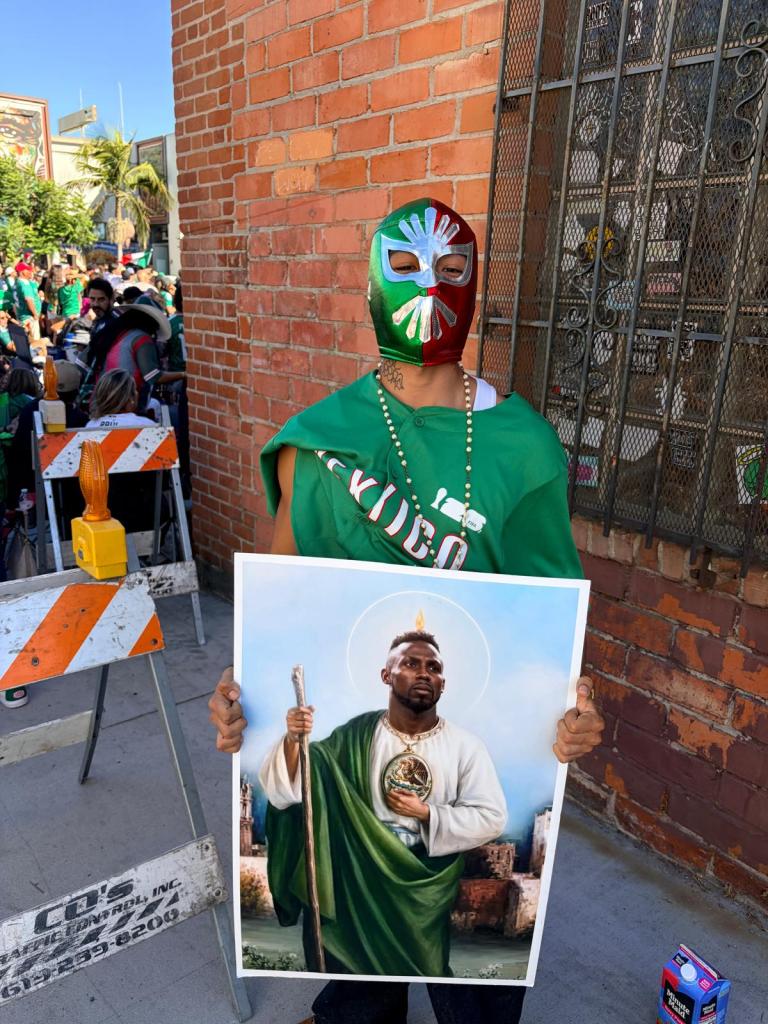 A man wearing a lucha libre mask and a green shirt holds a photo showing another man illustrated as a saint.