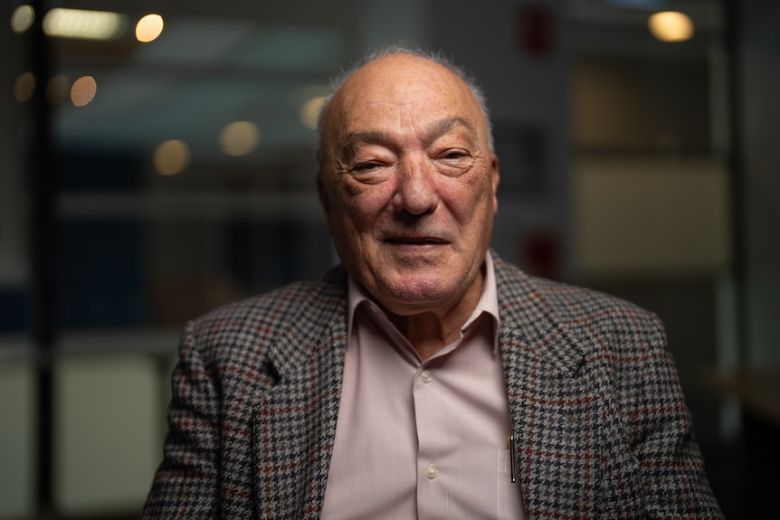 a bald man aged in his 60s or 70s wearing a tweed blazer over a pale pink collared shirt
