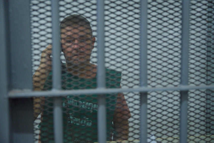 A man in a dark green prison outfit looks solemn as he holds a phone to his ear, behind metal bars and a thick security screen.