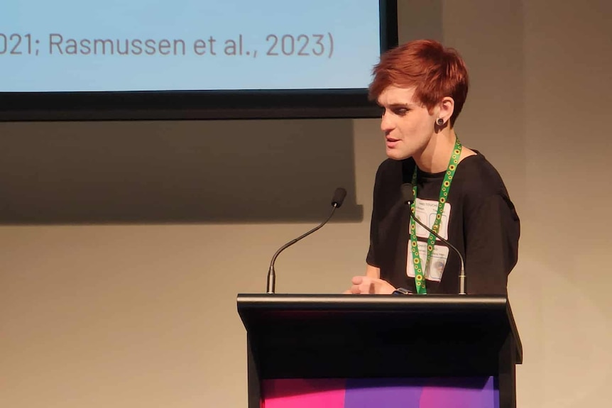 A speaker with short pink hair at a lecturn talks to the audience.