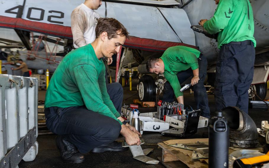 Sailors in green shirts working on aircraft parts in a ship’s hangar bay.