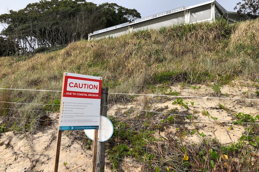 A 'caution' sign in front of a badly eroded dune with a safe in the background.