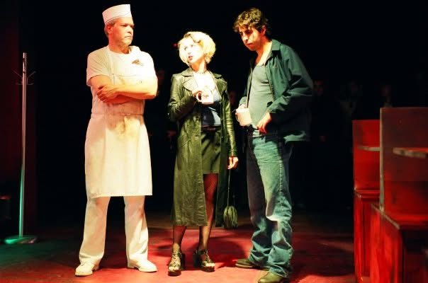 A man in a white cook's outfit stands left, with a woman in dark coat and heels and a short dress center. A man in jeans and a cloth windbreaker stands right. There are diner booths to their right.