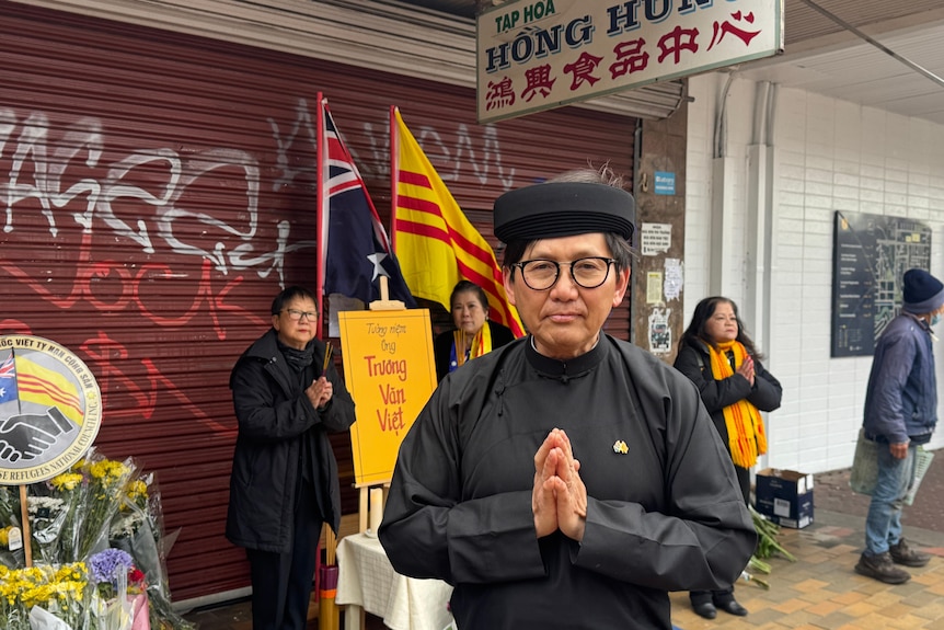 A man in a black headdress and clothes stands with his palms together in prayer near women doing the same outside a shop.